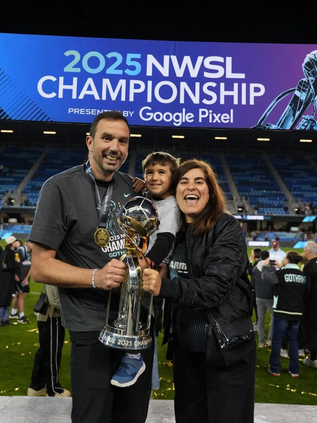 Family celebrating the 2025 NWSL Championship, holding the trophy with a stadium screen in the background.