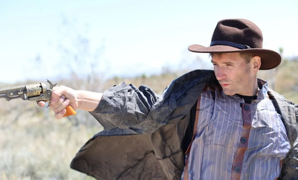 Man in cowboy hat and costume holding a revolver in a desert setting.