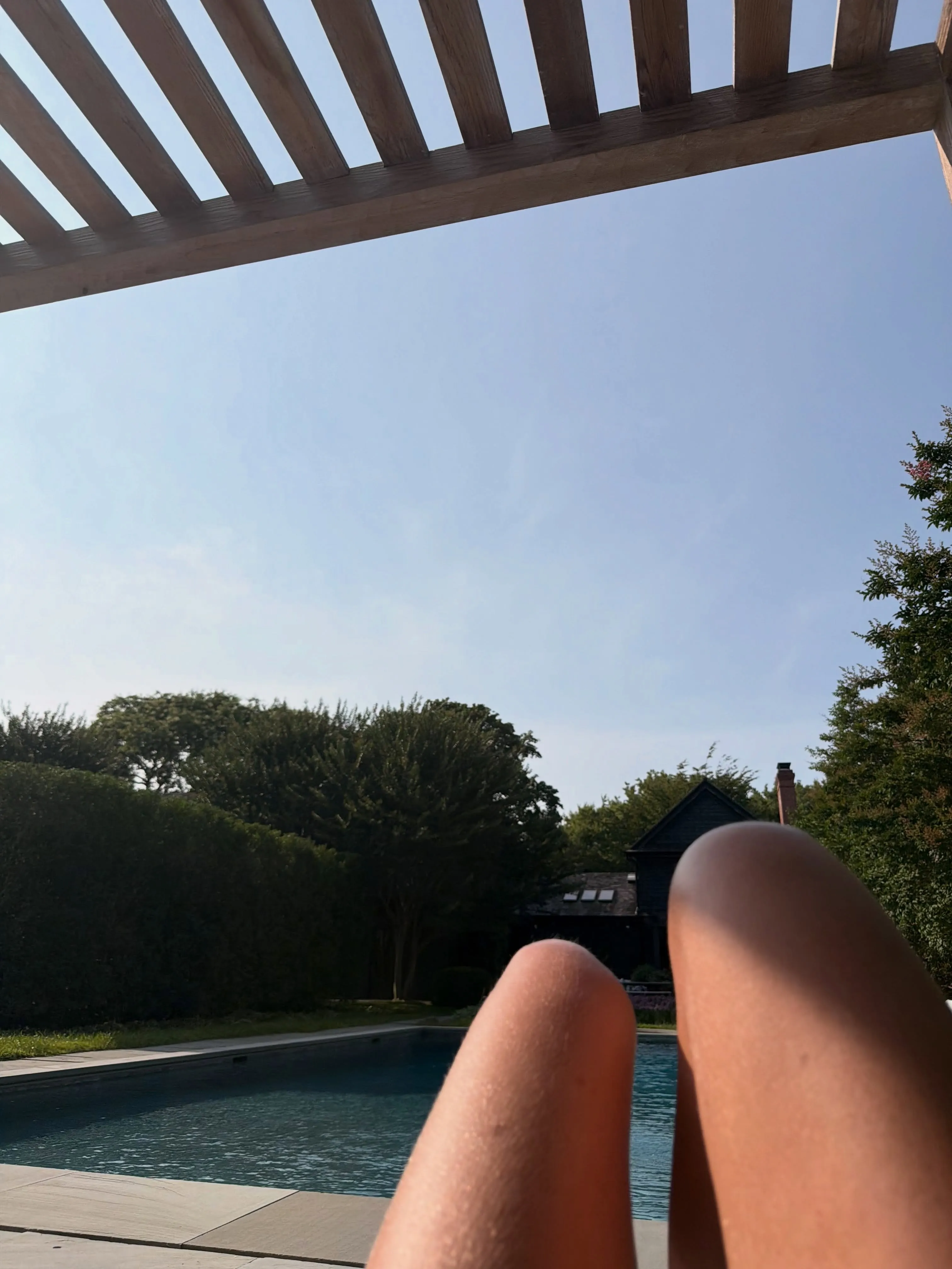 A first-person view of legs lounging by a swimming pool under a wooden trellis, with trees and a house in the background.