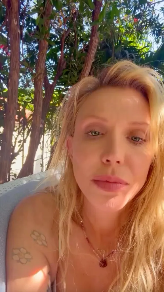 A blonde woman with green eyes and a nude pink lip looking directly at the camera, with trees in the background.