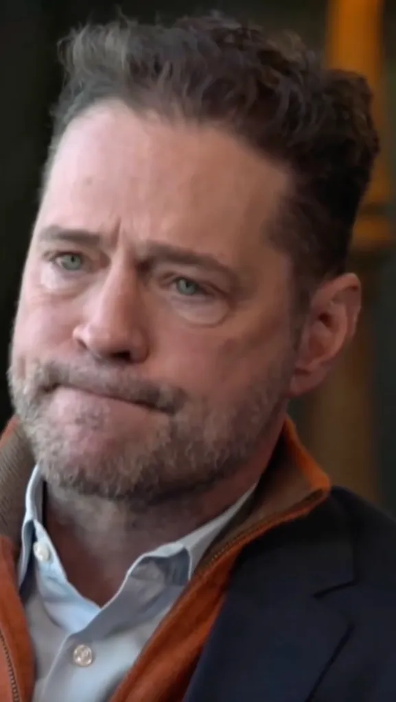 A man with a beard and light blue eyes wearing a blue collared shirt with an orange and dark blue jacket.