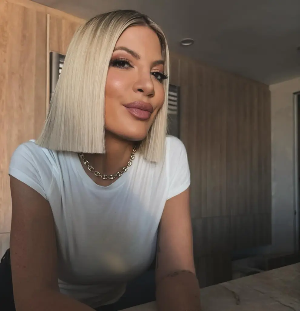 A woman with straight blonde hair and a white t-shirt looks at the camera.