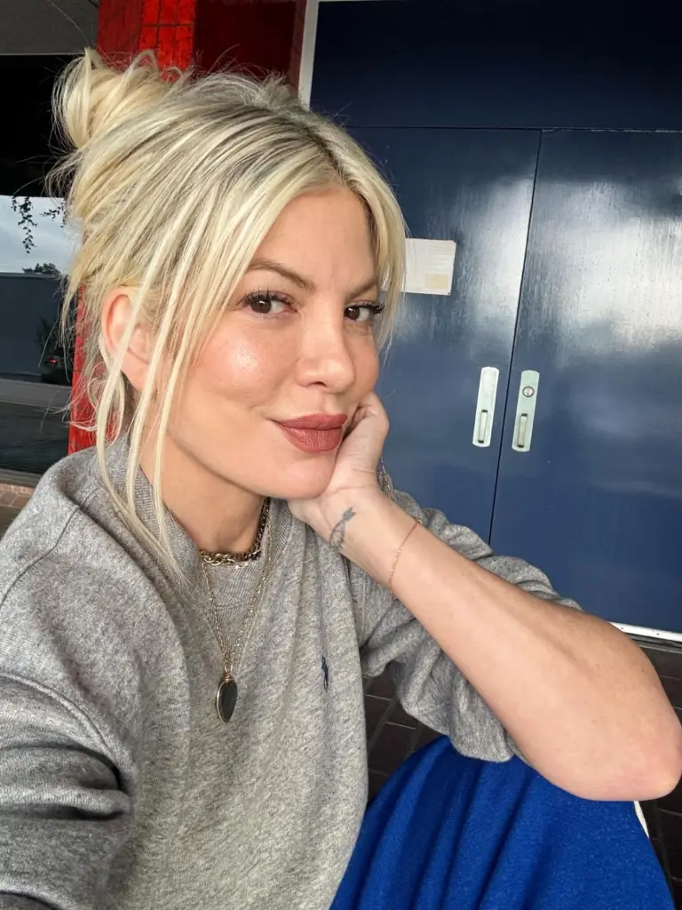 A blonde woman with her hair in a messy bun, wearing a grey sweatshirt and blue pants, leans on her hand.