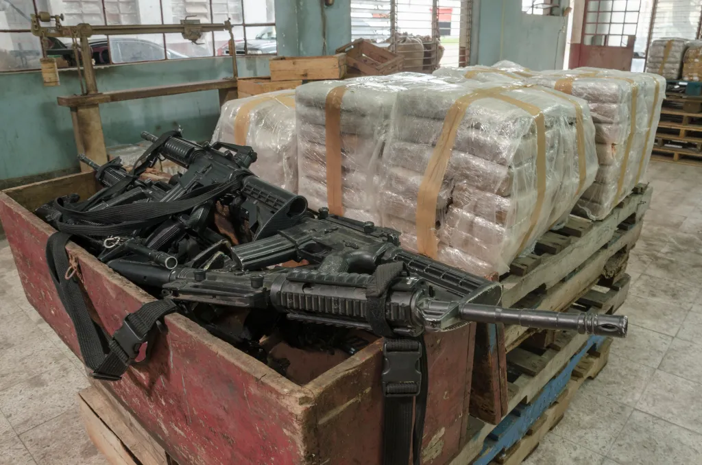 A crate filled with firearms next to pallets of cocaine bricks wrapped in plastic.
