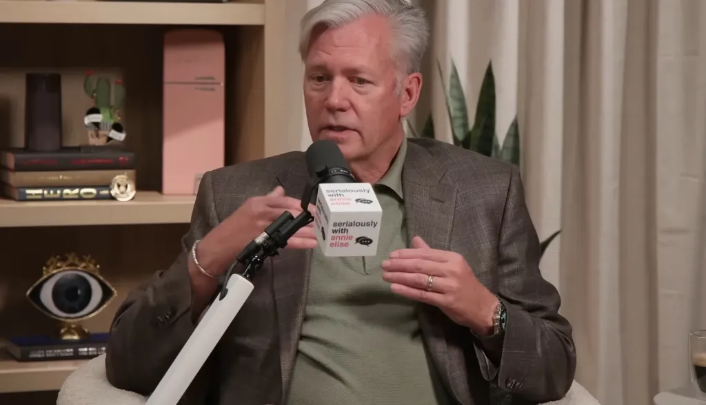 A man in a suit jacket gestures while speaking into a microphone.