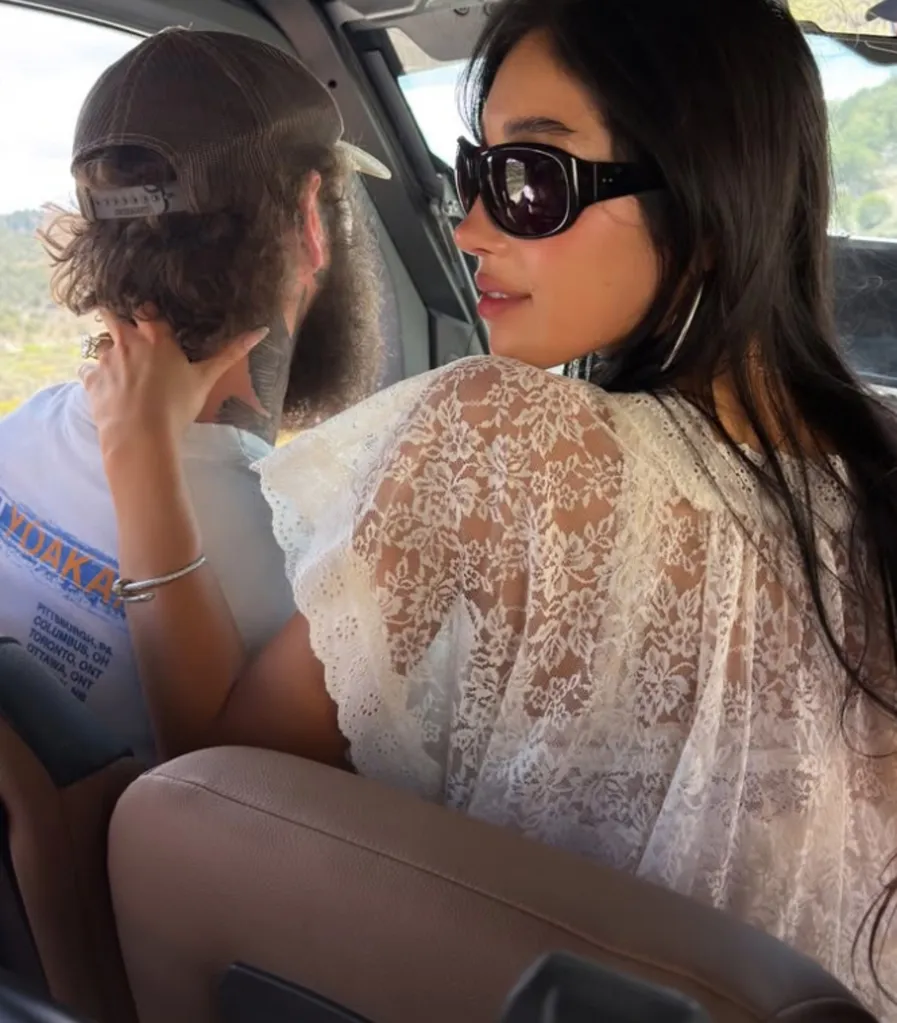A woman in sunglasses and a white lace top looks over her shoulder while a man in a baseball cap drives.