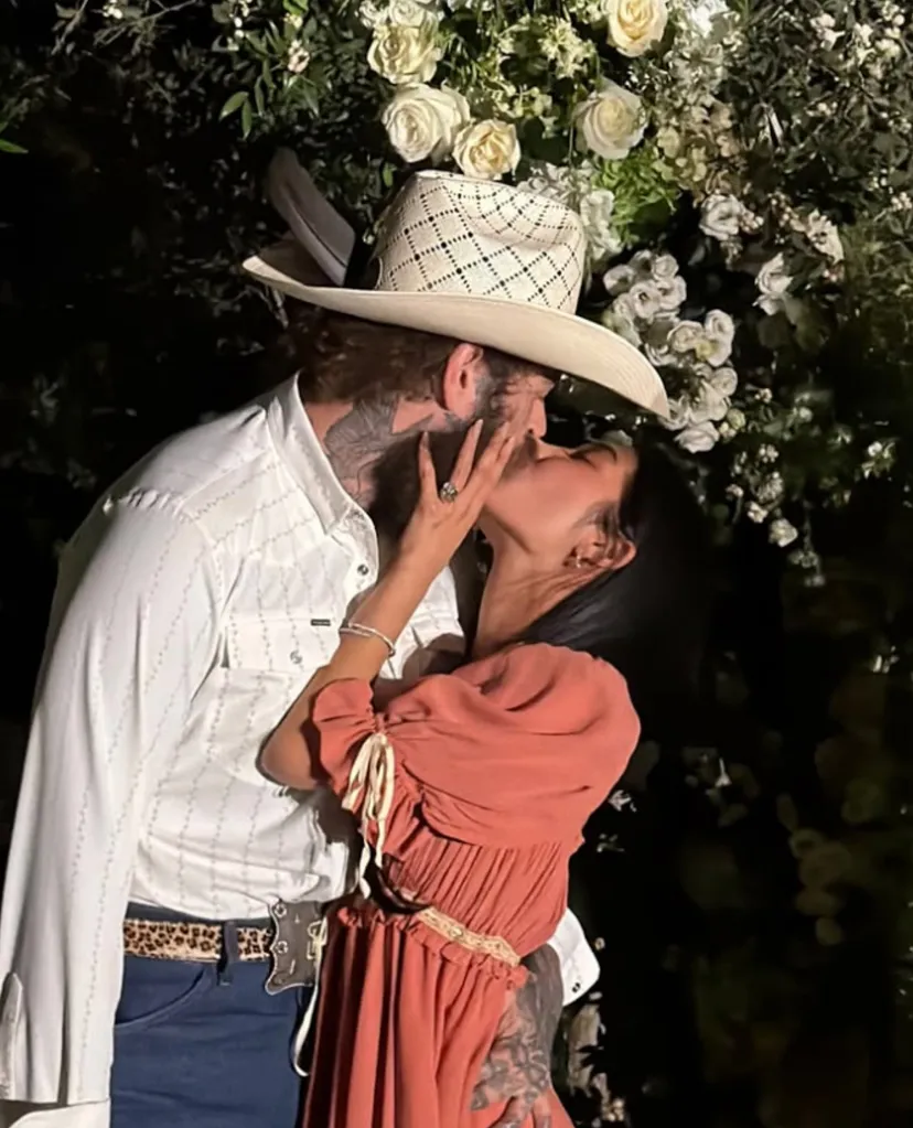 A man in a cowboy hat and a woman in an orange dress kissing, with a floral arrangement in the background.