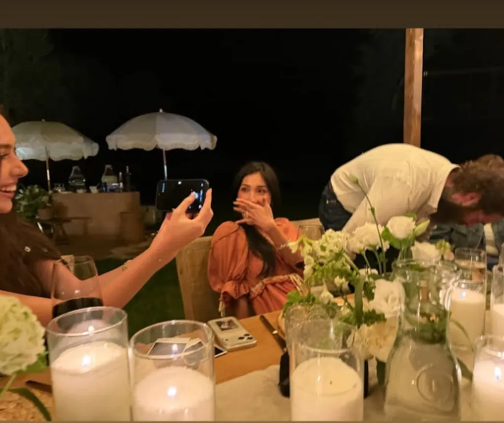 A woman holding a phone records a woman at a table covering her mouth, as a man bends over in the background.