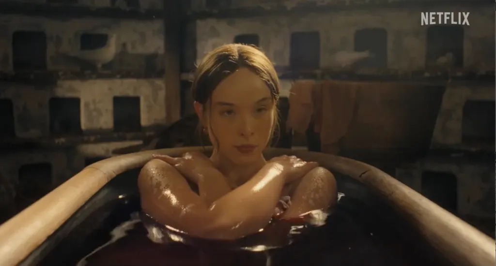 A young woman with auburn hair sits in a wooden tub filled with dark water, her arms crossed and glistening with moisture.