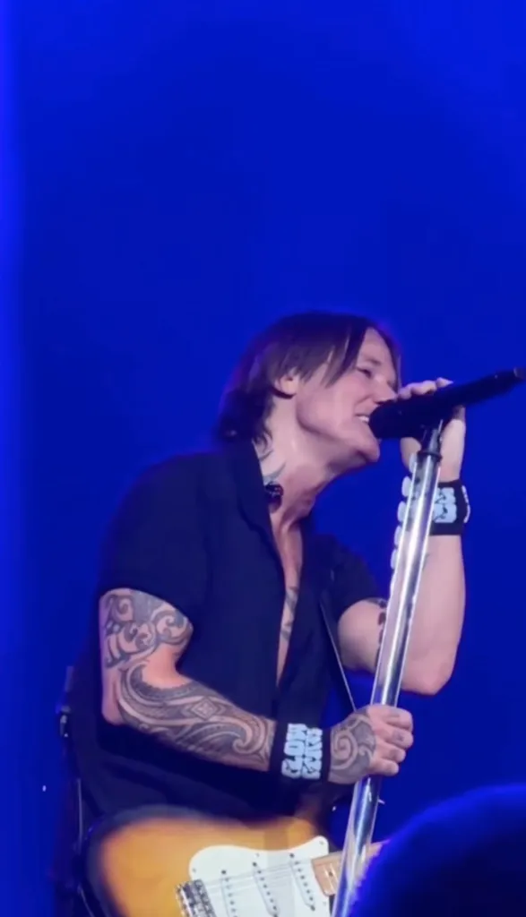A male singer with tattoos on his arms and neck performs with a microphone and guitar against a blue background.