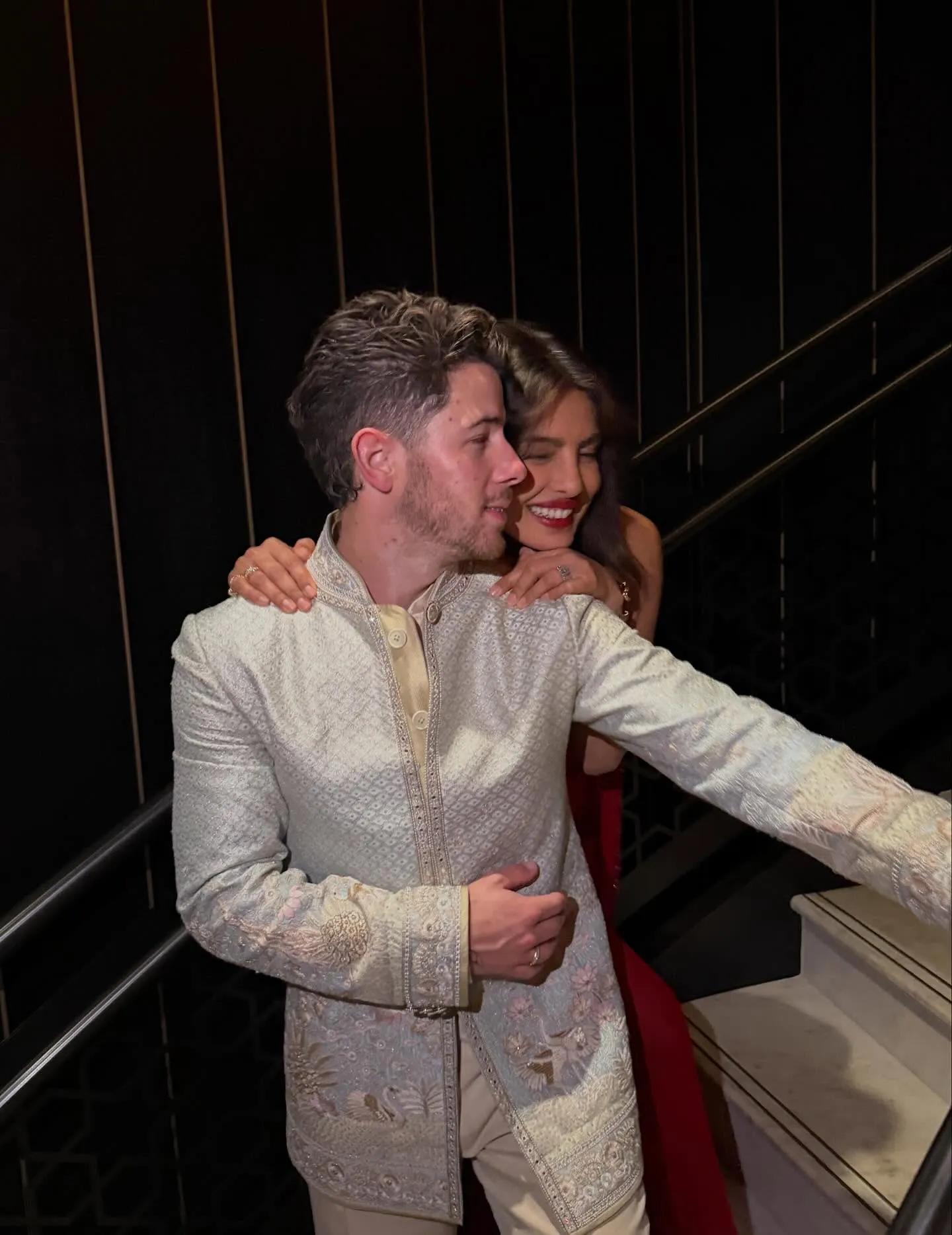 Nick Jonas wearing a traditional Indian outfit with Priyanka Chopra smiling over his shoulder, her hands on him.