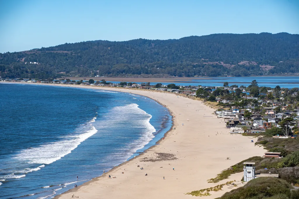 Stinson Beach, a long sandy beach with waves breaking on the shore, bordered by a town with houses and green hills in the background.