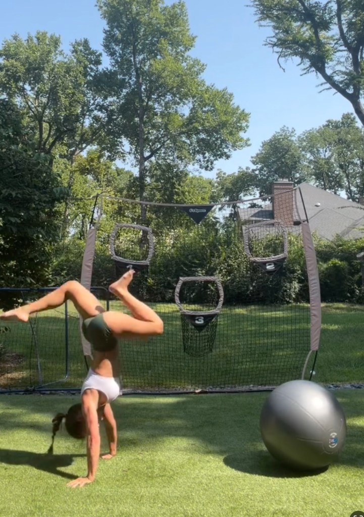 A young woman doing a handstand on green turf with a sports netting and a large stability ball in the background.