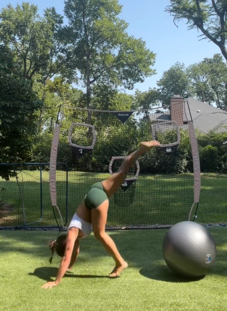 Woman in athletic wear doing a downward dog pose with one leg raised on a green turf.