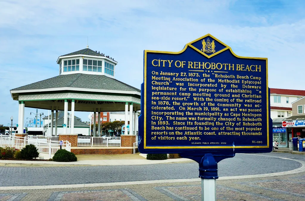 A historical sign for the City of Rehoboth Beach, Delaware, with a gazebo and buildings in the background.
