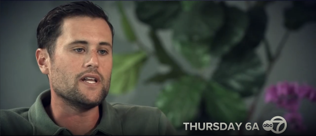 A young man with dark hair and a beard, wearing a green polo shirt, speaks to the camera.