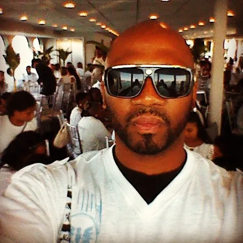 A man with a goatee and sunglasses taking a selfie in a crowded room.