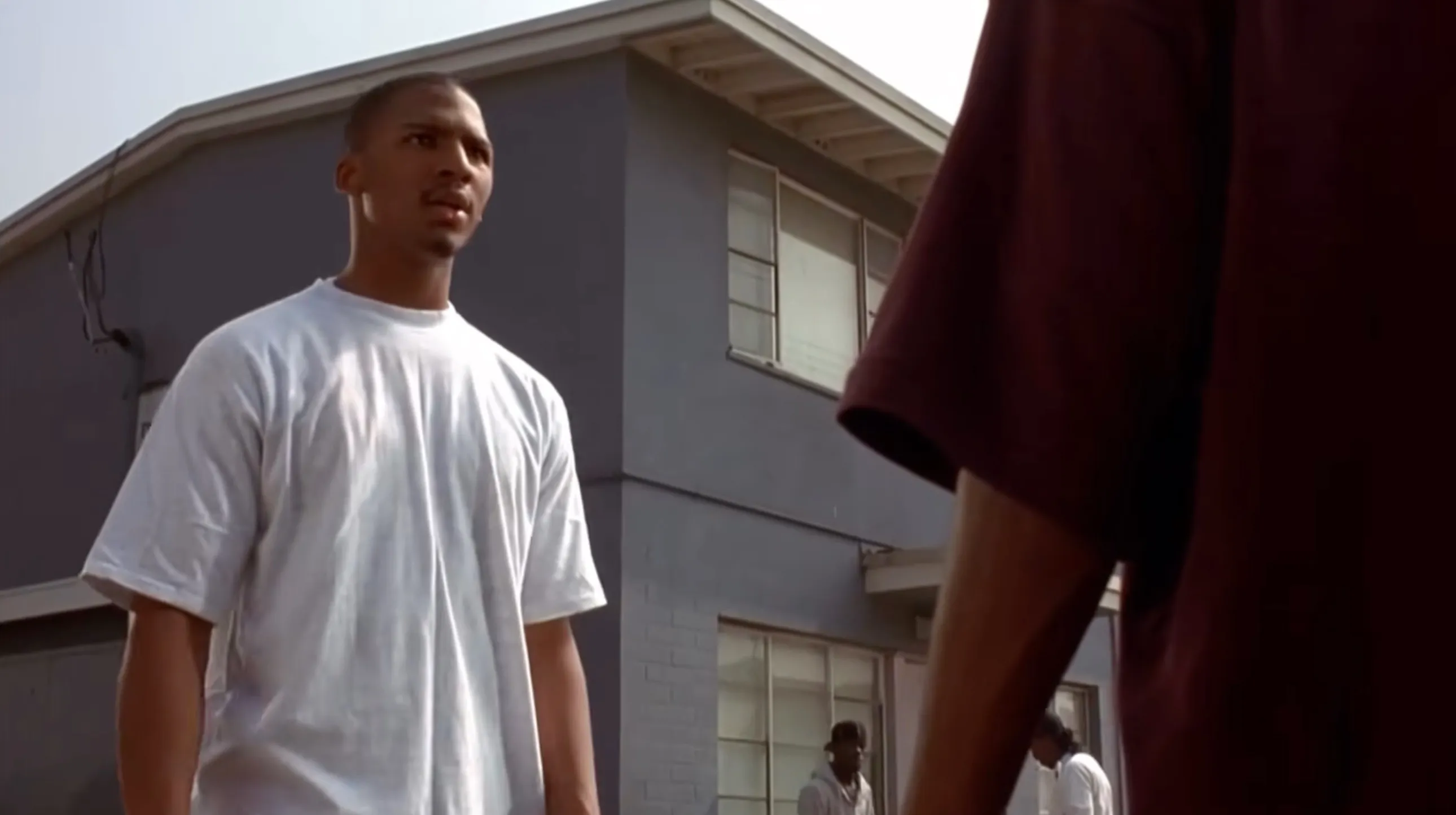 A young Black man in a white t-shirt looks forward, with part of another person's red t-shirt visible in the foreground.
