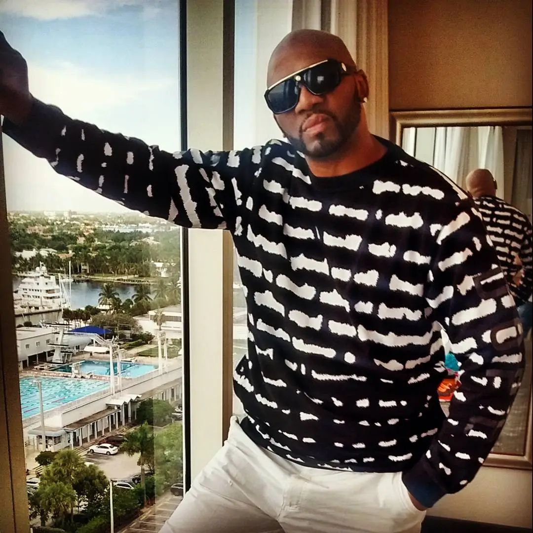 A man in a black and white patterned shirt poses next to a window overlooking a city with a marina.
