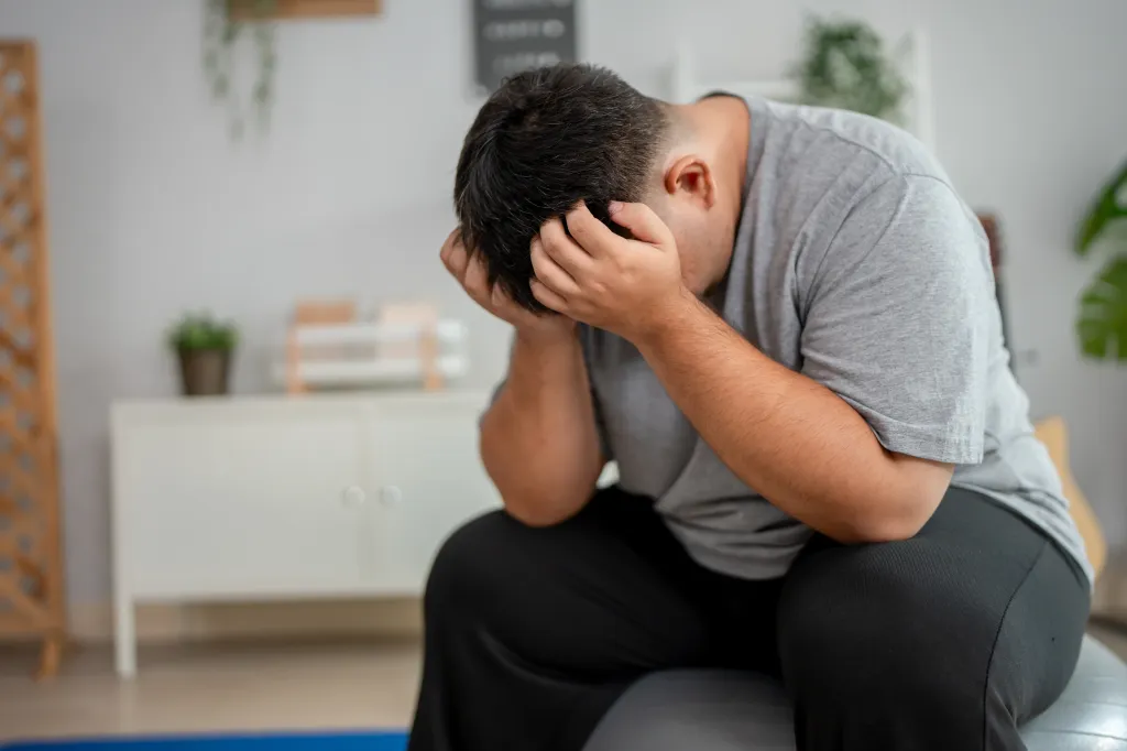 Exhausted plus-size man sitting on an exercise ball with his head in his hands.