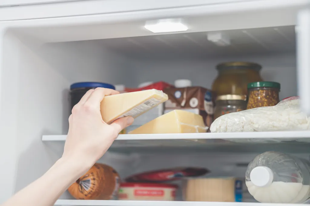 A person's hand reaching into a refrigerator to take out a wedge of Parmigiano cheese.