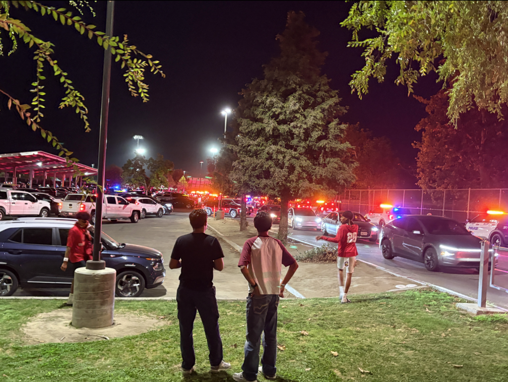 People gathered at night in a parking lot, watching emergency vehicles with flashing lights.