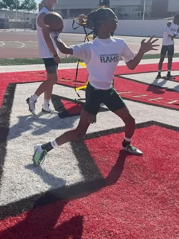 A boy in a white Rams shirt playing football on a red turf field.