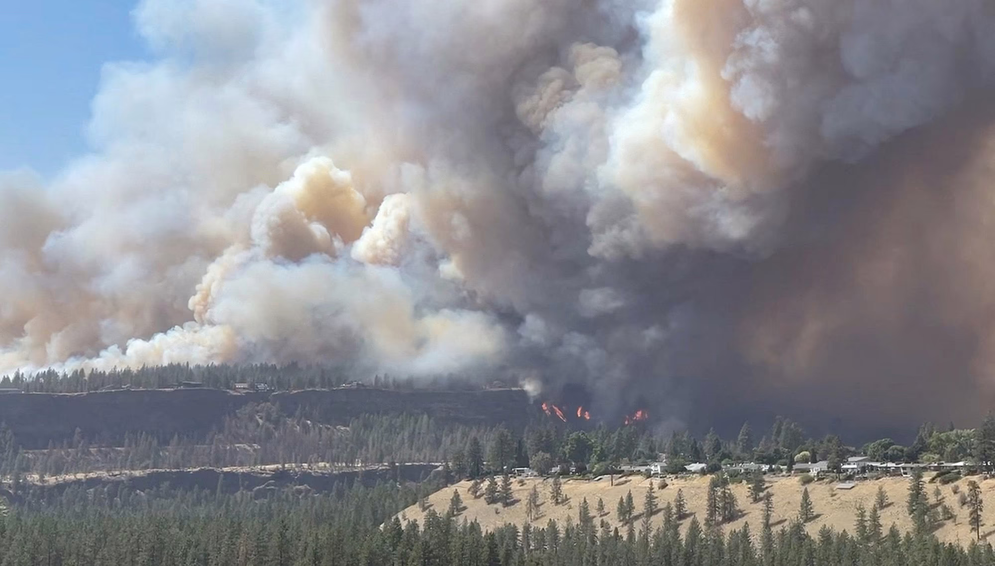 Large smoke plumes rise from a wildfire seen on a distant bluff.