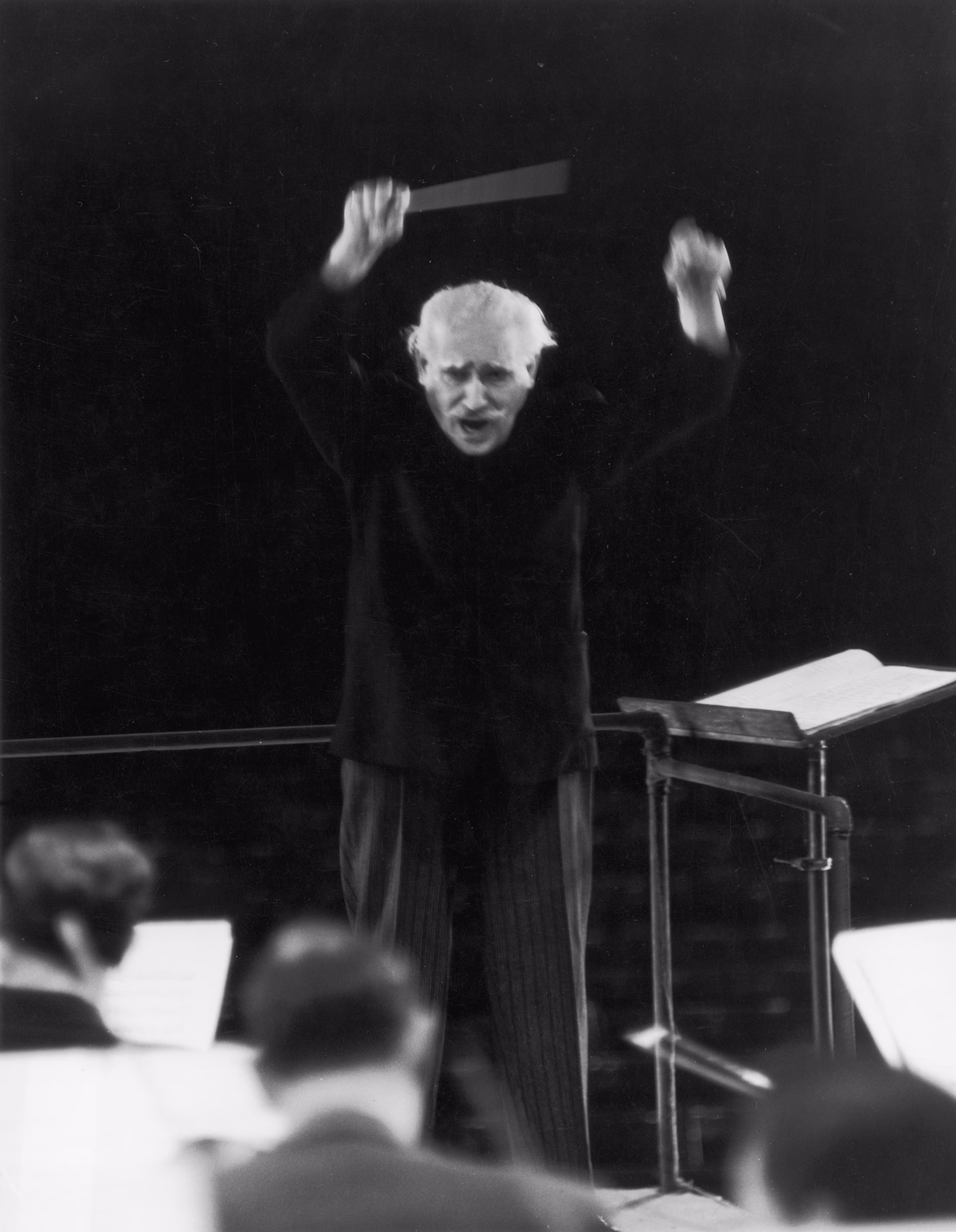black-and-white photo of mustachioed, white-haired man with arms raised above head, baton in right hand, score to his left, with orchestra members in foreground