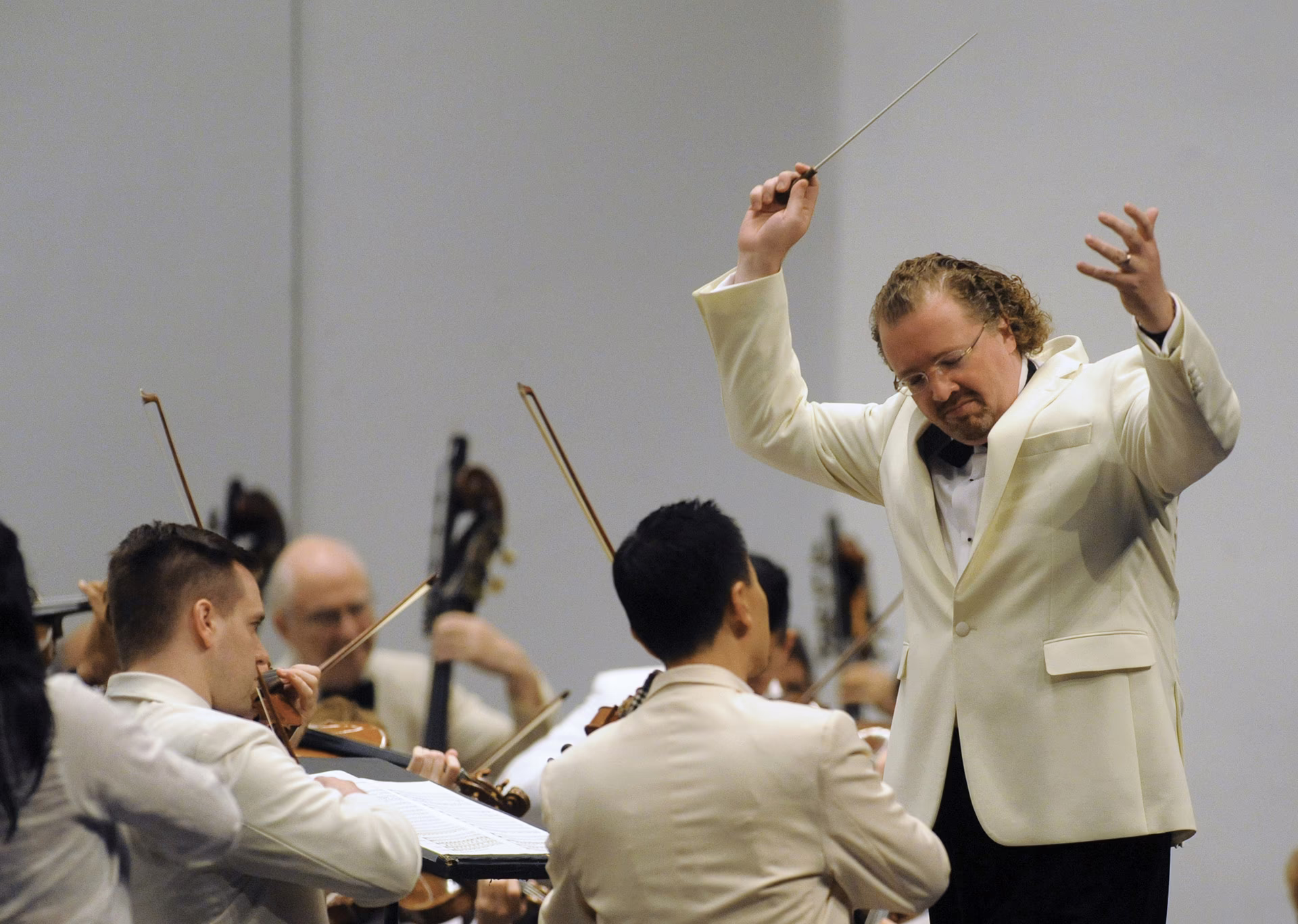 color photo of man in white jacket and black tie and pants with arms raised and eyes closed standing before string section