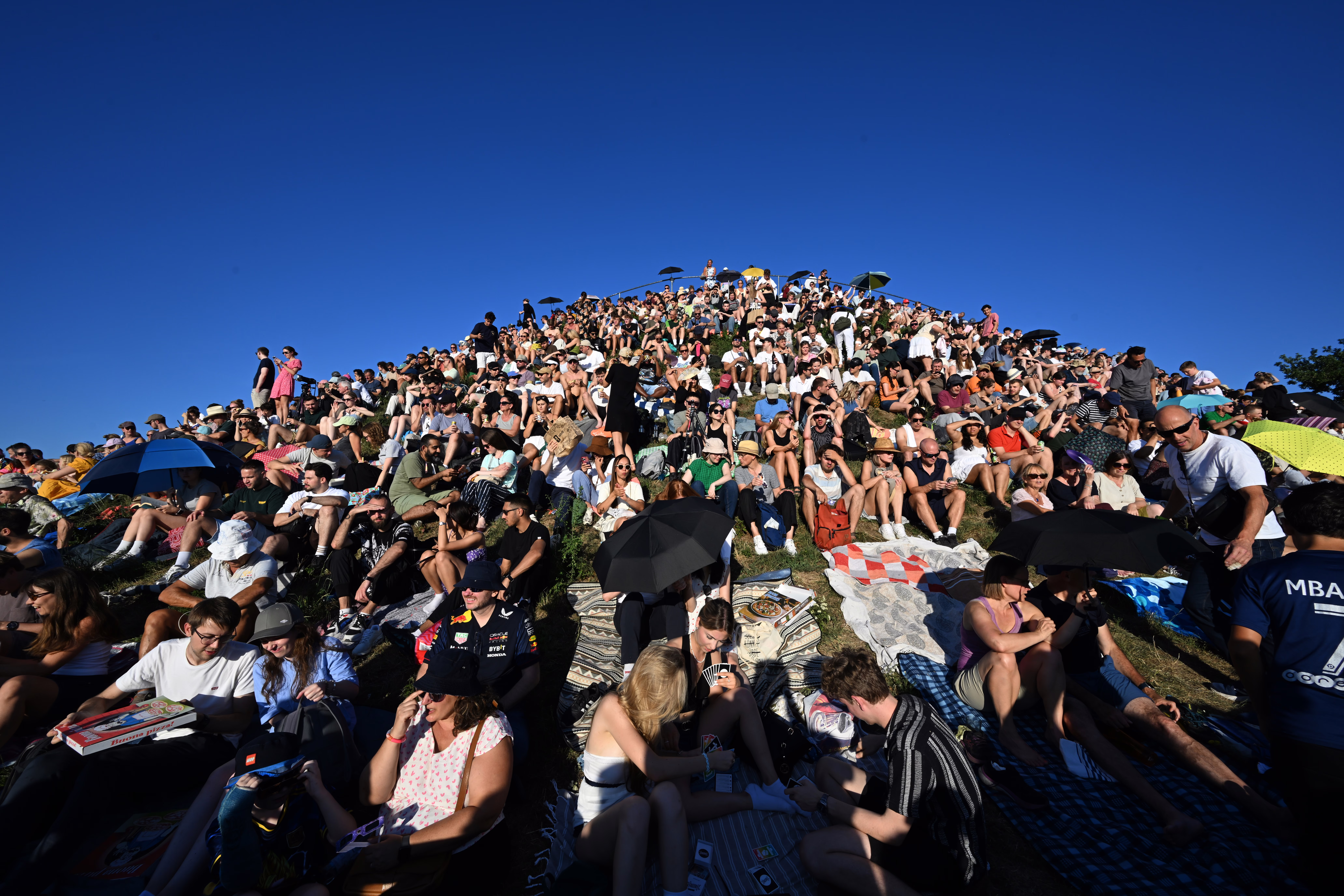 Many people sit on a hill in a park, under a sunny sky.
