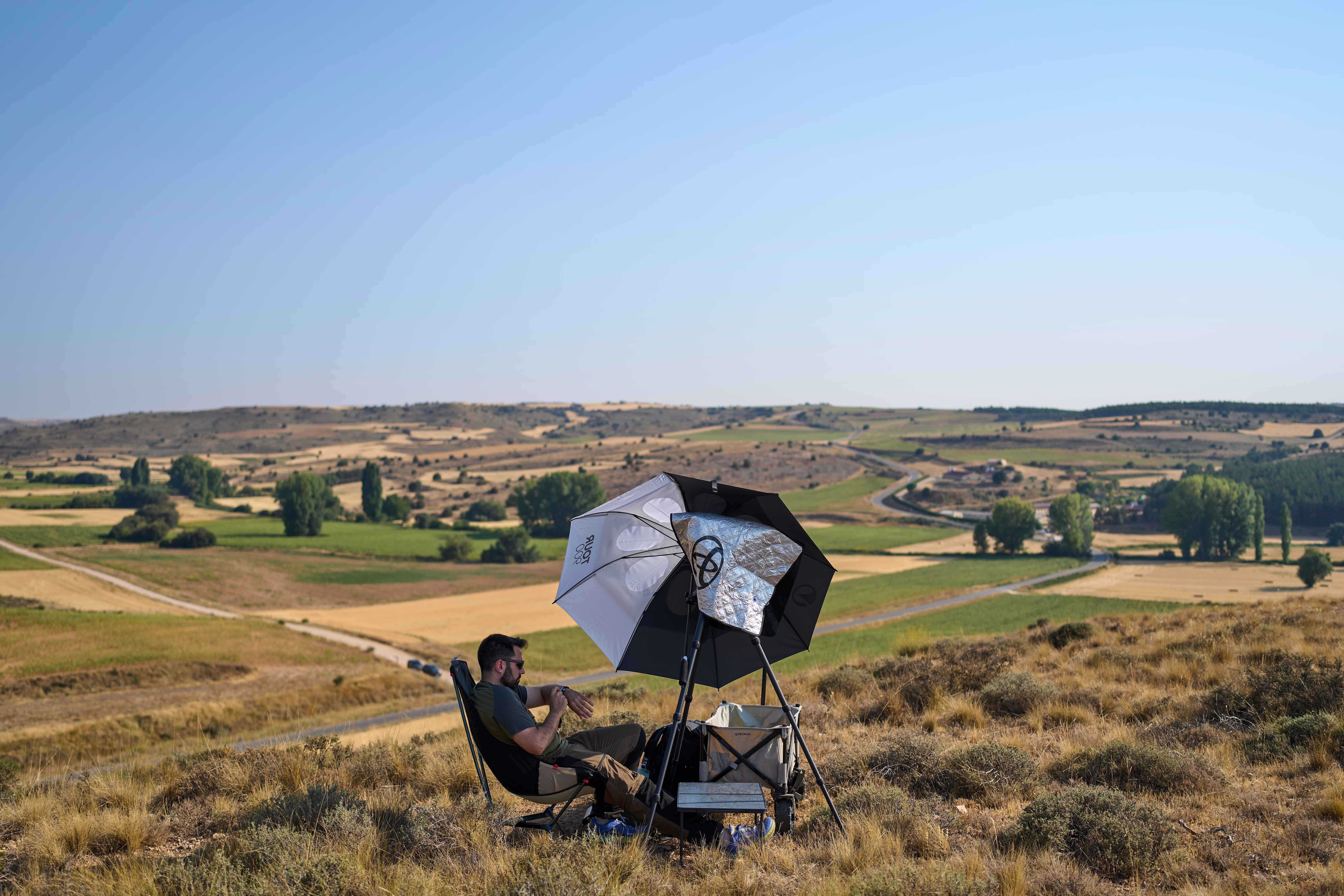 A person sits on a hillside, beneath an umbrella, with some equipment.