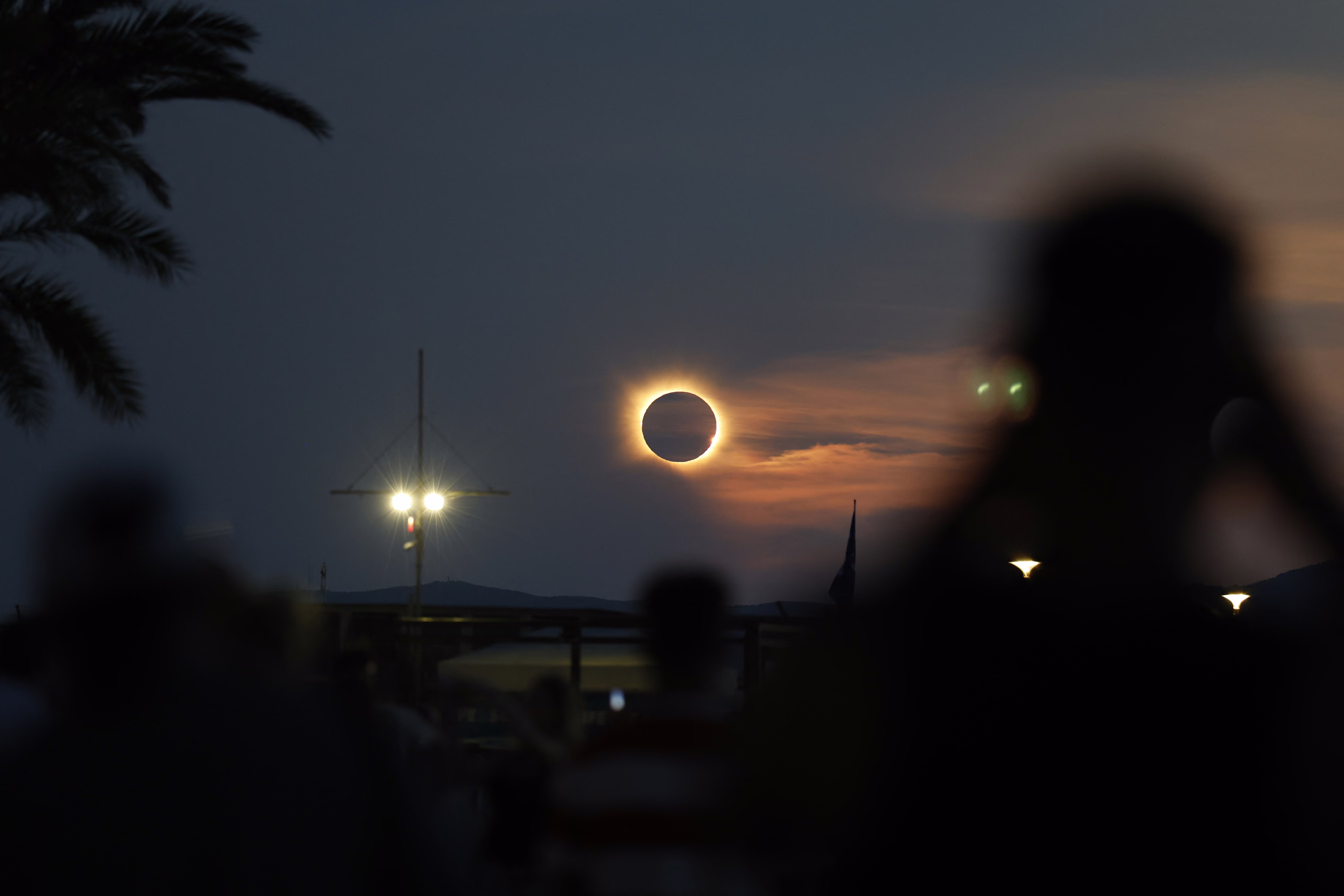 People look at a total eclipse, seen behind light clouds, near the horizon.