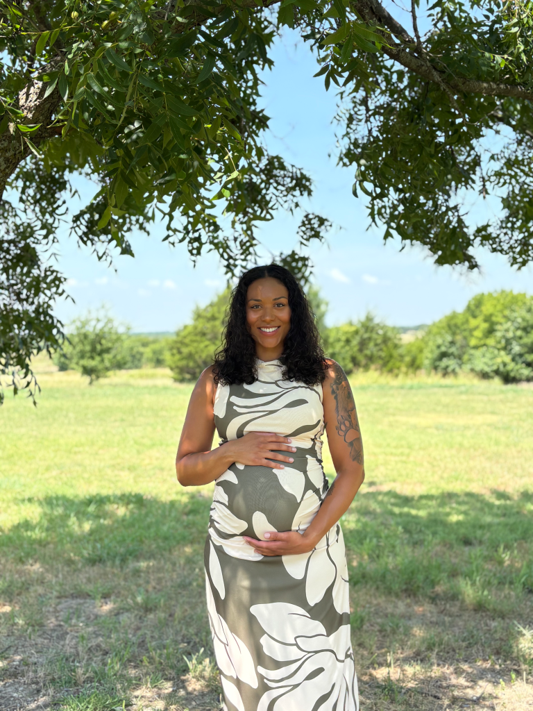 McKenna West smiles while holding her pregnant belly under a tree.