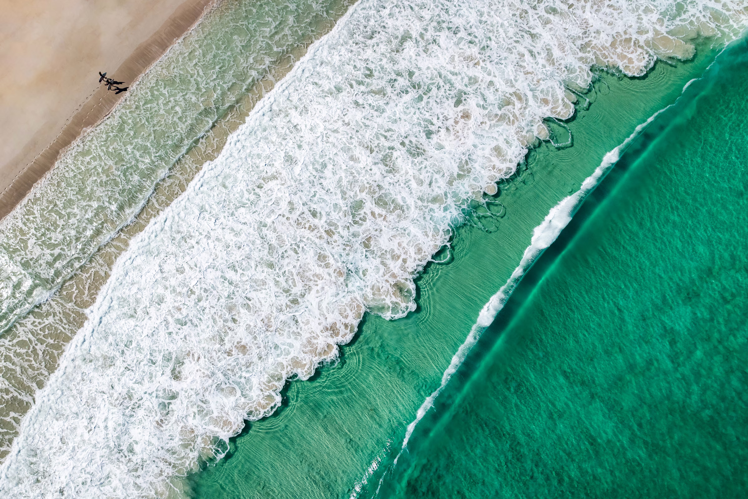 An aerial view of waves crashing on a beach, with clear green water behind them.