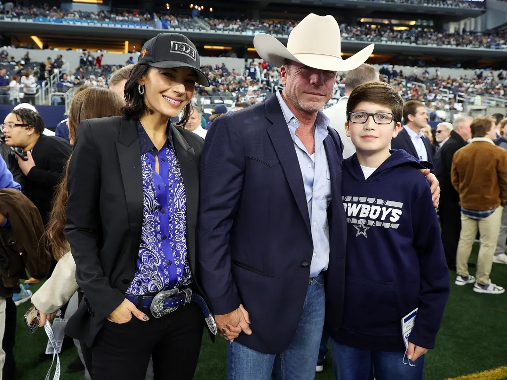 Nicole Muirbrook, in a blue and white patterned shirt, black blazer, black jeans with buckled belt, black baseball cap, and Taylor Sheridan, in a cream cowboy hat, navy blue blazer, blue shirt, blue jeans, and Gus Sheridan in a Dallas Cowboys blue hooded sweatshirt and blue jeans, n the sidelines of an American football game.