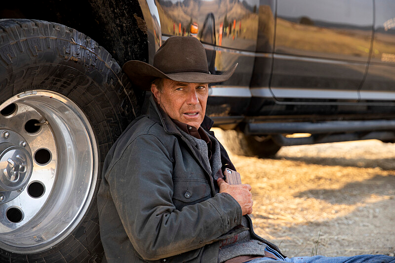 Kevin Costner as John Dutton in a dark jacket and brown cowboy hat and jeans, leans against a truck tire, holding a phone to his chest.