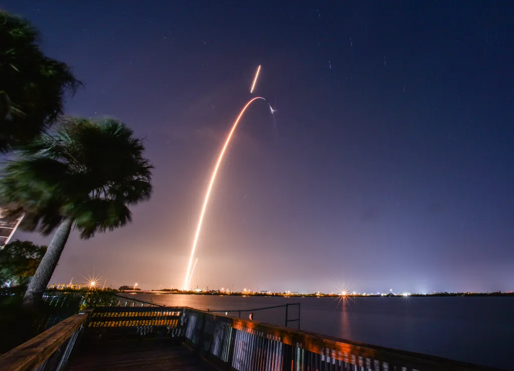 A long-exposure photo showing the launch of a SpaceX Falcon 9 rocket and its booster landing at night, with palm trees and a body of water in the foreground.