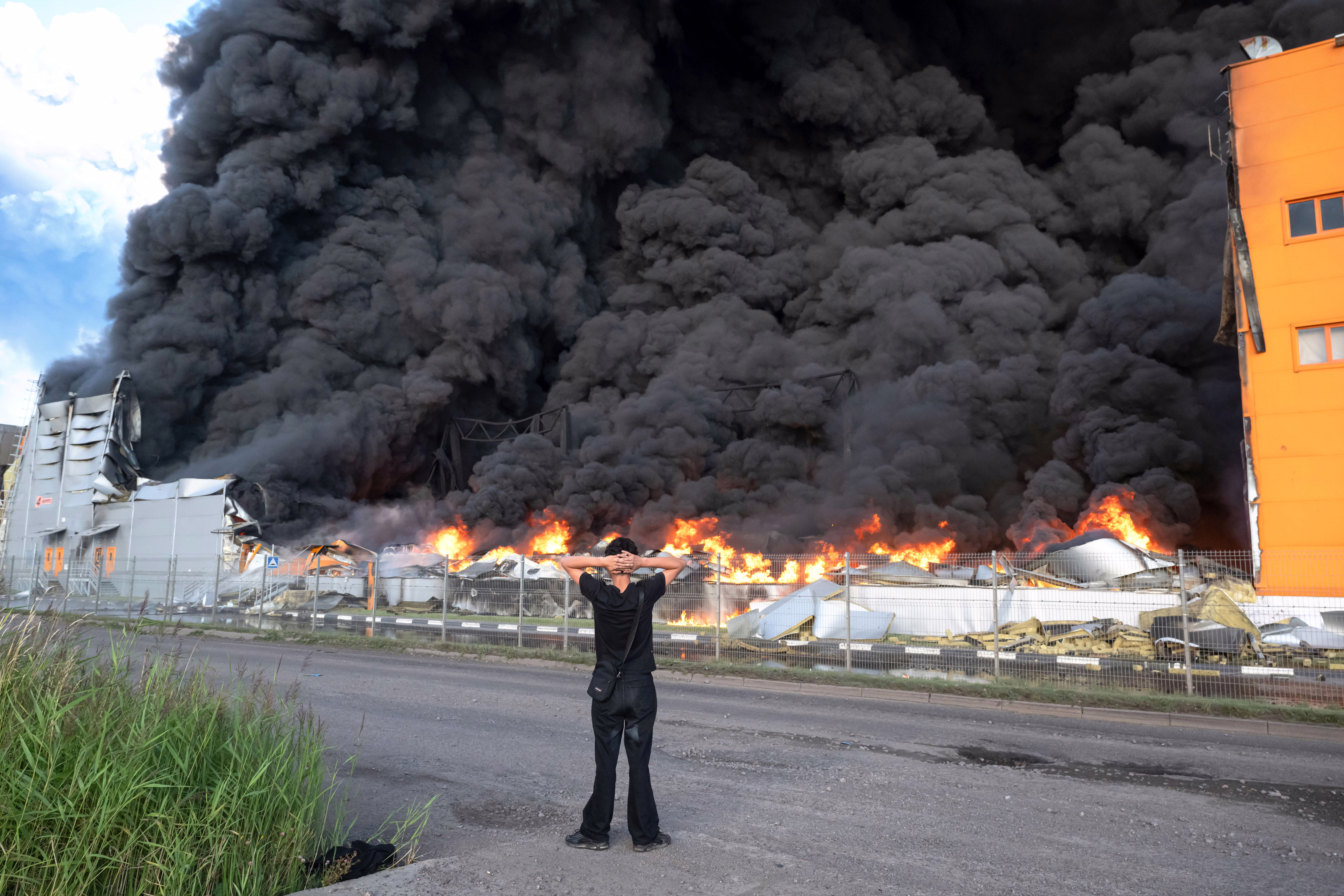 A man stands on a roadside, looking at at thick black smoke rising from a burning warehouse.