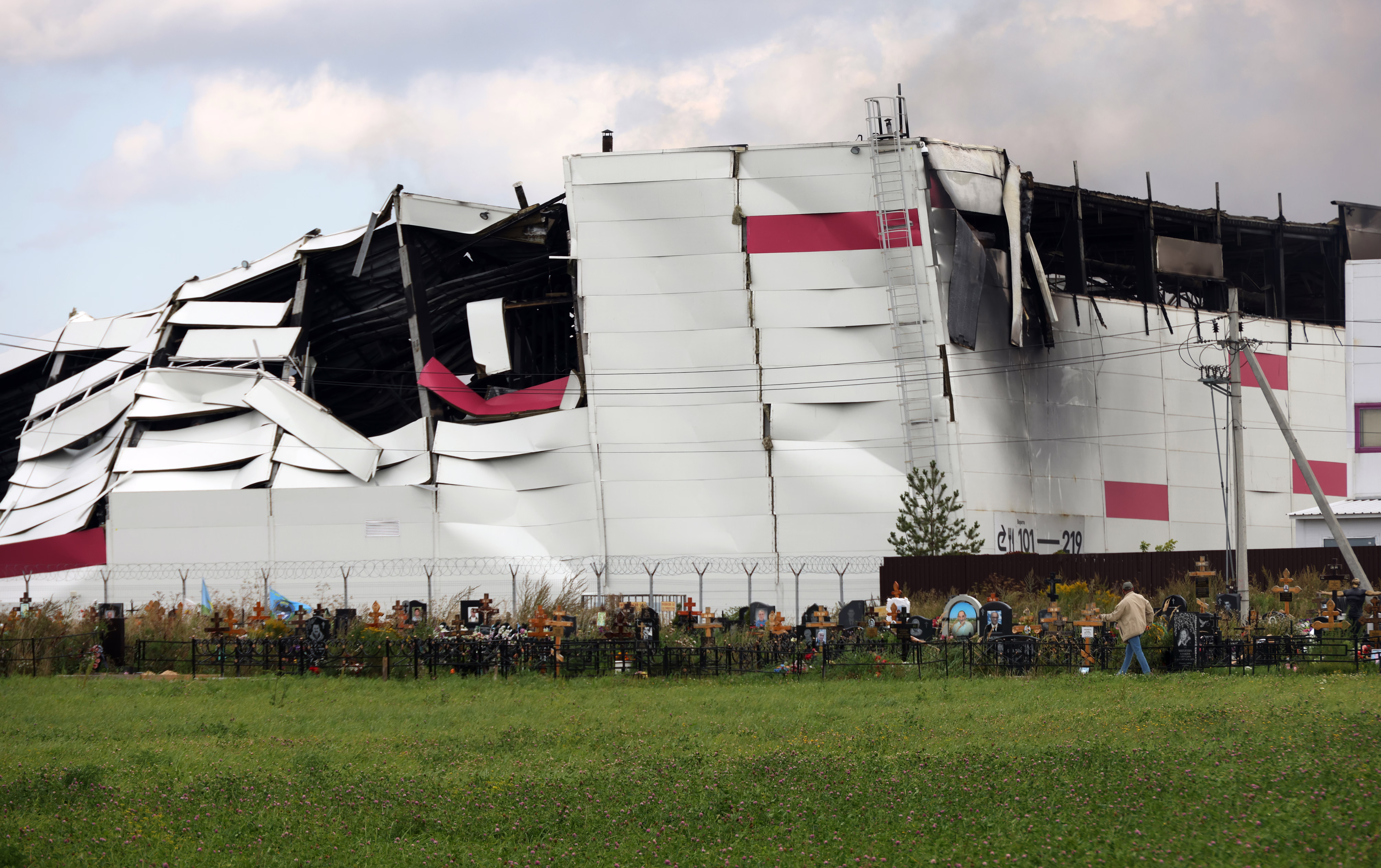 A person walks past gravestones that stand near the ruins of a burned-out warehouse in the background.