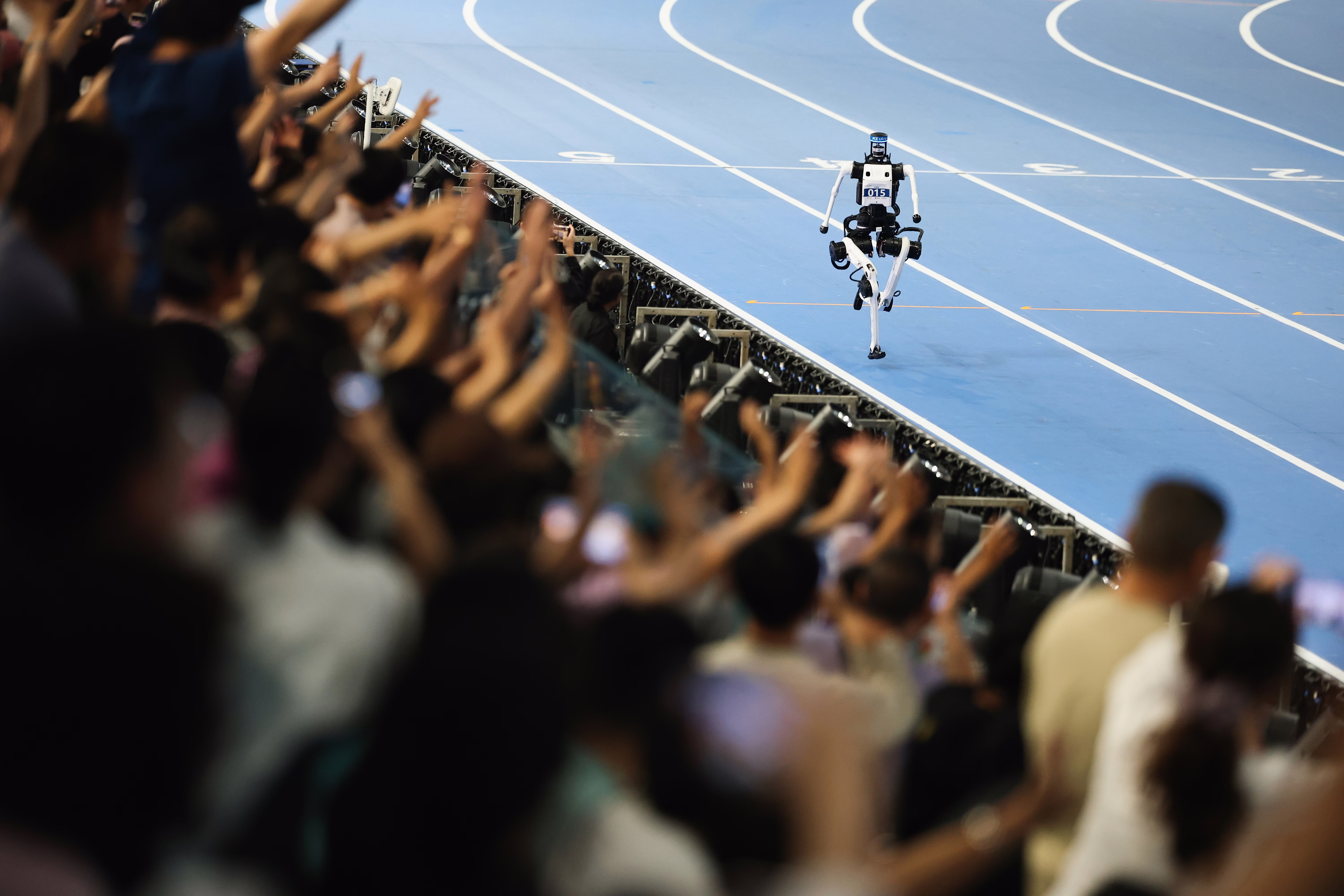 A crowd cheers as a humanoid robot runs past them on a track.