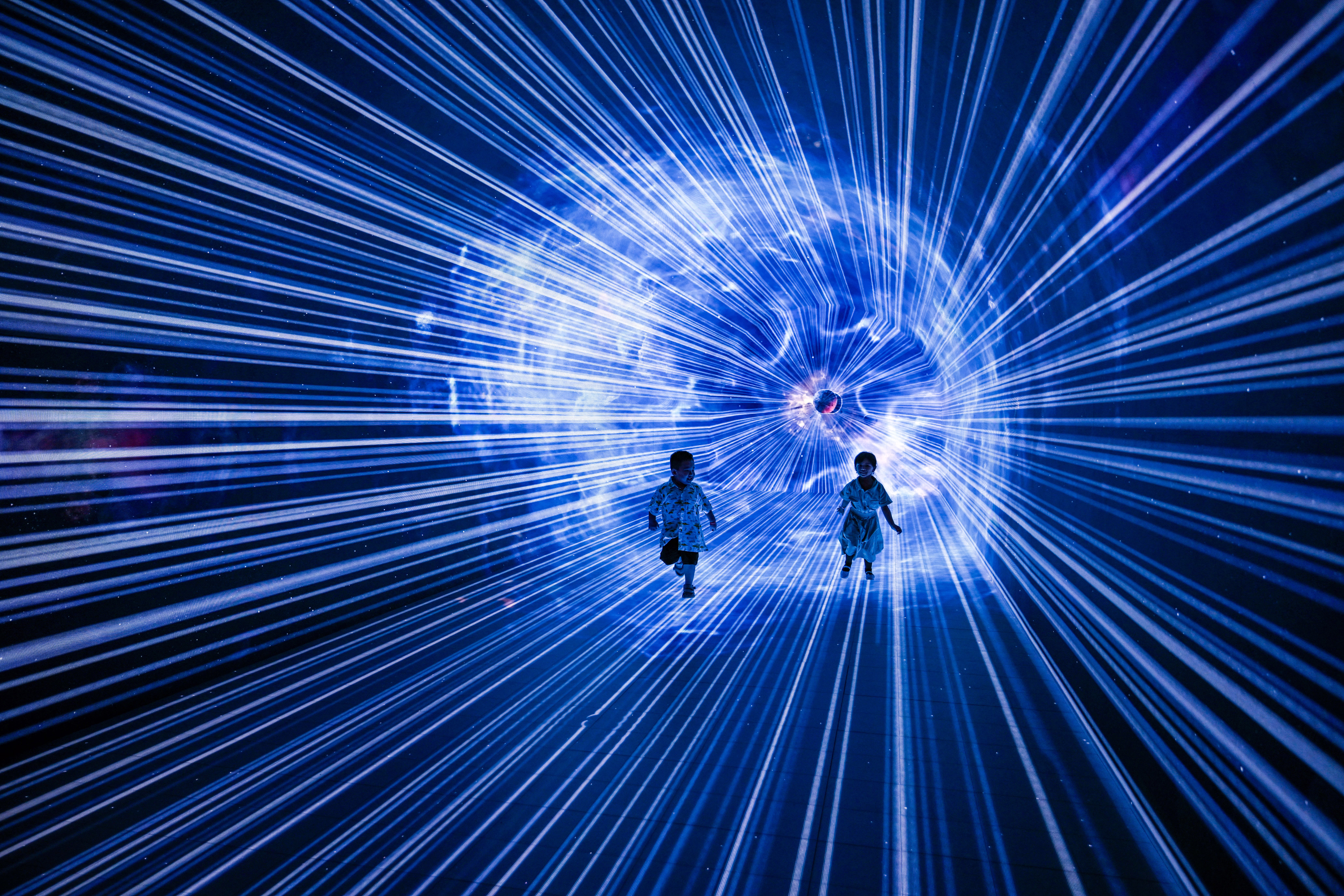 Two young children run inside an exhibit space with walls illuminated by bright striped projections.