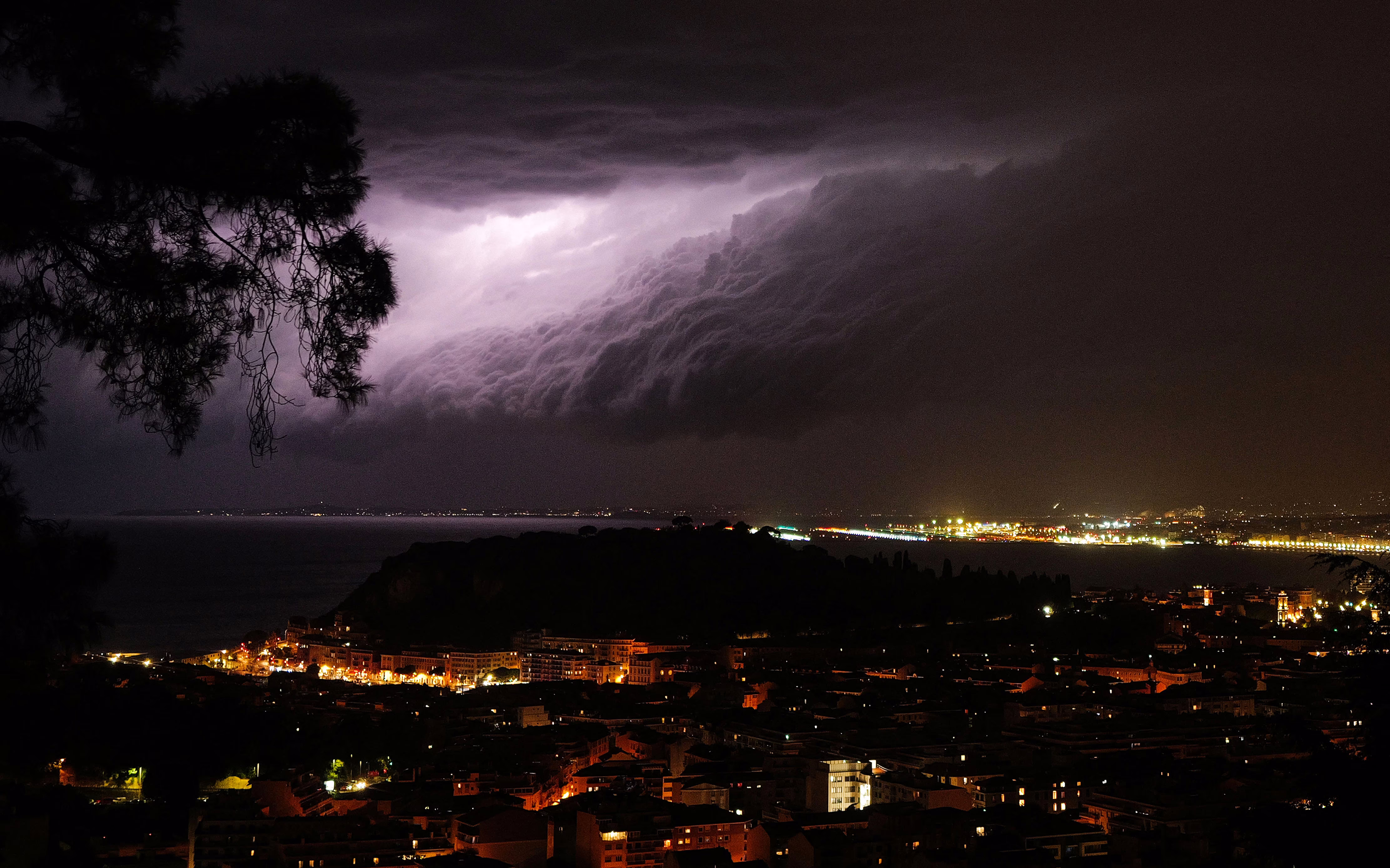 Lightning strikes in the clouds over a coastal city at night.