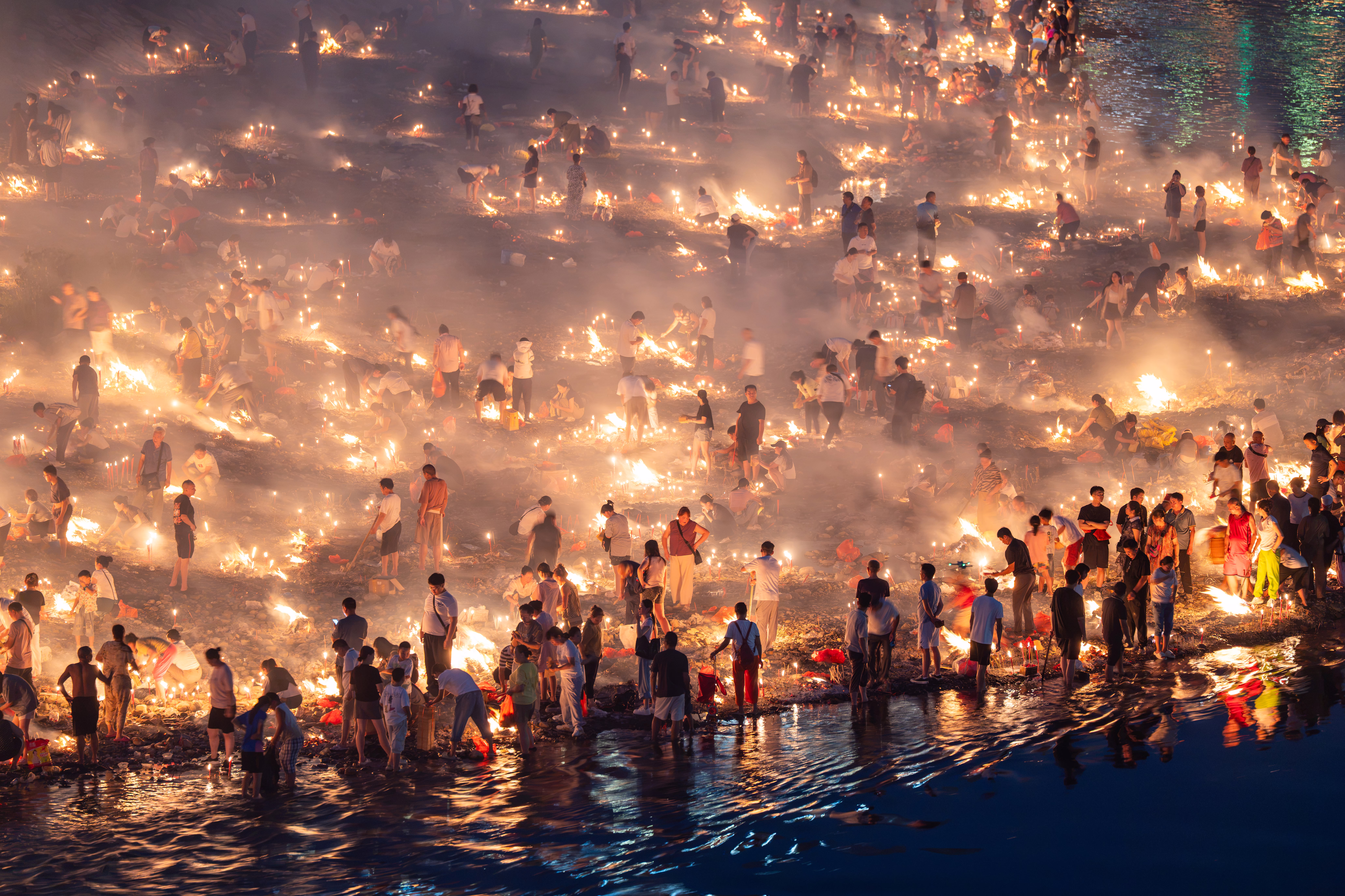 Dozens of groups of people burn paper offerings on a riverbank.