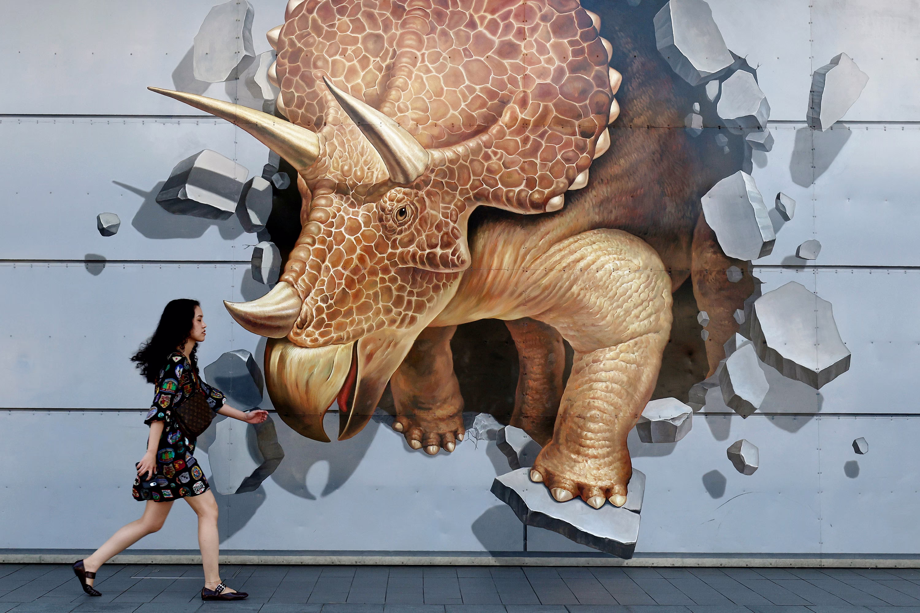 A woman walks past a giant dinosaur mural, depicting a triceratops bursting through a wall.
