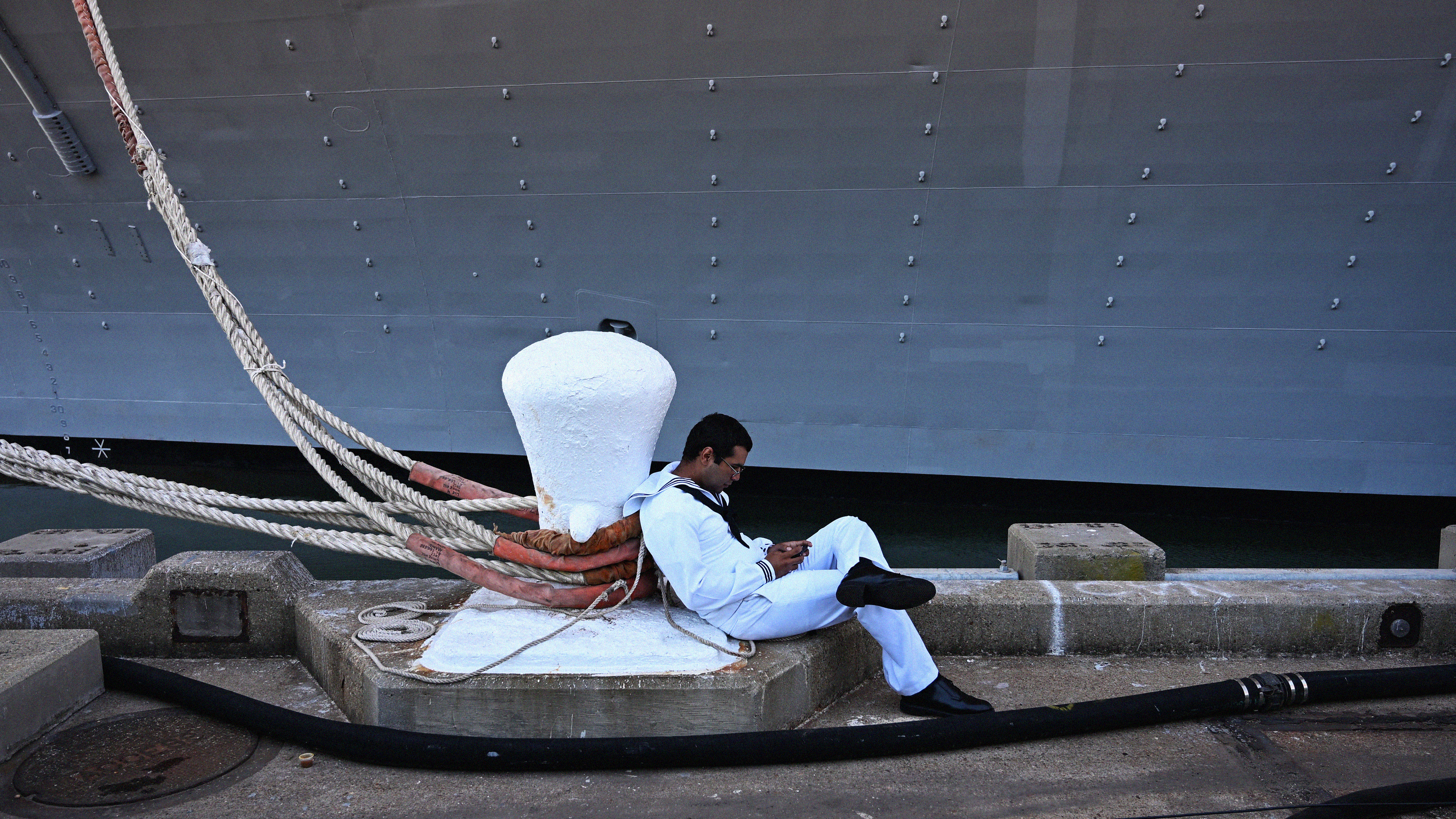 A sailor in a white uniform leans against a white bollard on a dock in front of a ship