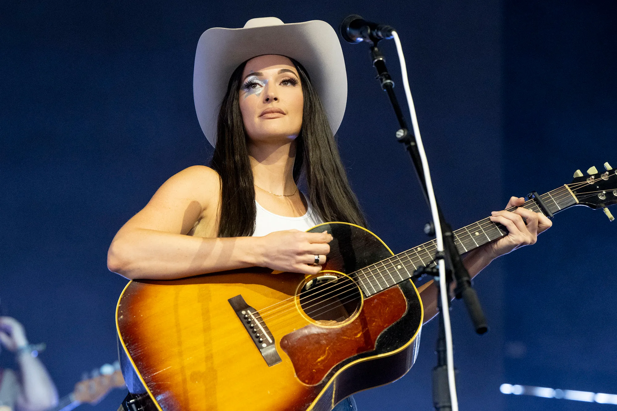 Kacey Musgraves performing on stage at Coachella, wearing a white cowboy hat and playing an acoustic guitar.
