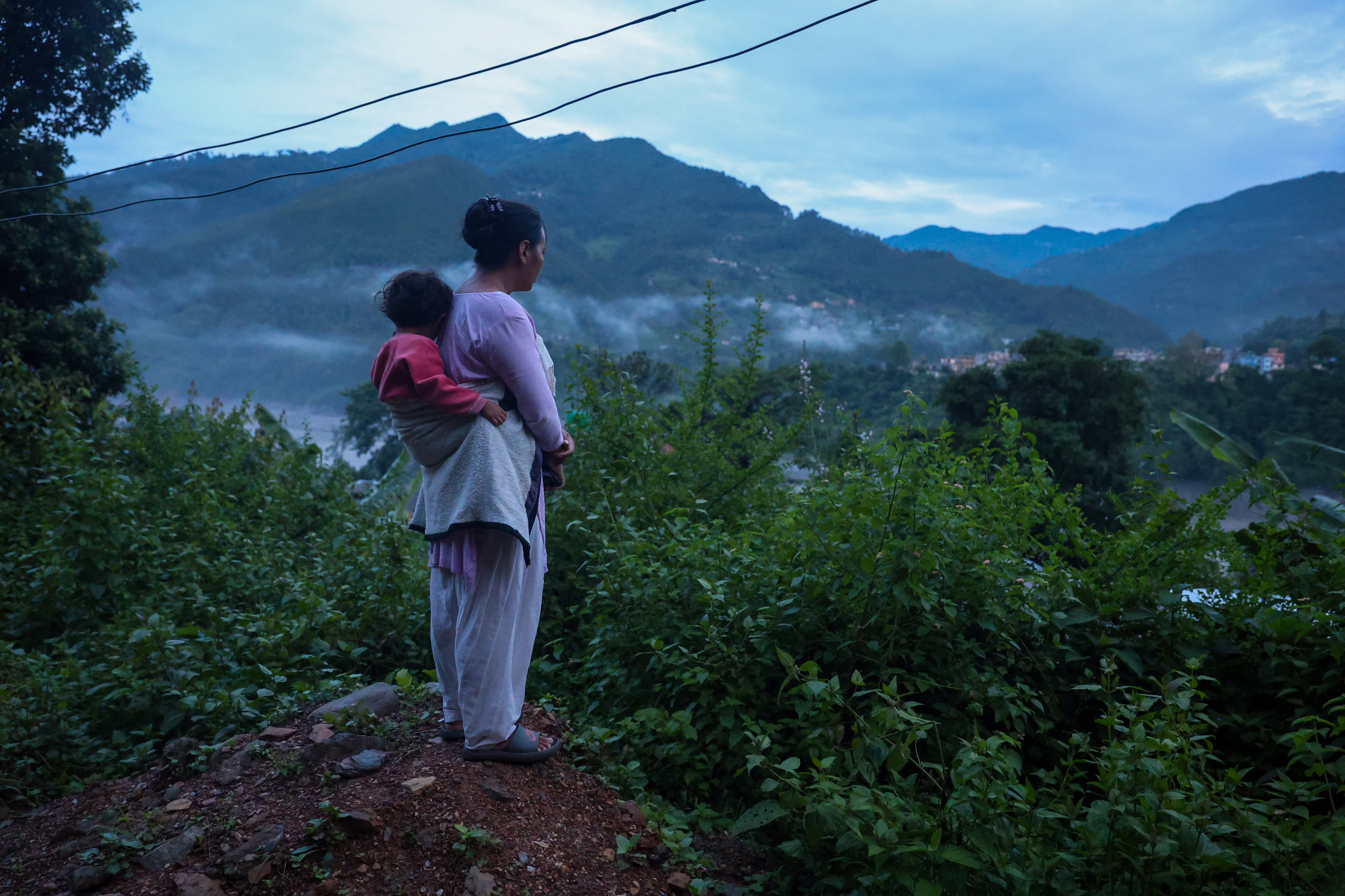 A woman, carrying a small child on her back, looks out over a river valley.