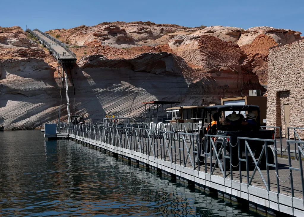 A dock extends over the water of Lake Powell towards rocky banks, with a long ramp leading up to the top of the cliff.