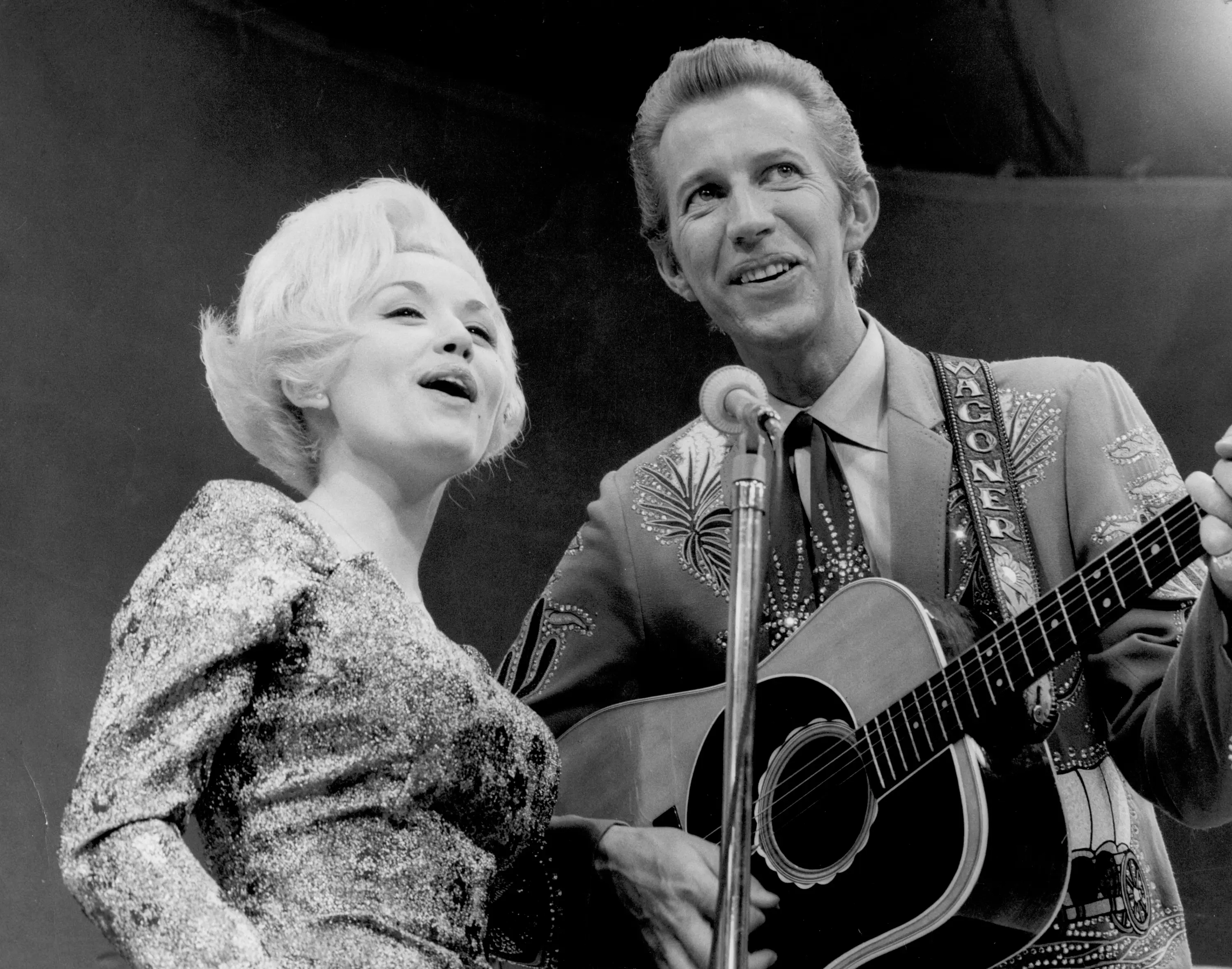 Porter Wagoner in a Nudie Suit, playing guitar alongside Dolly Parton singing into a microphone.